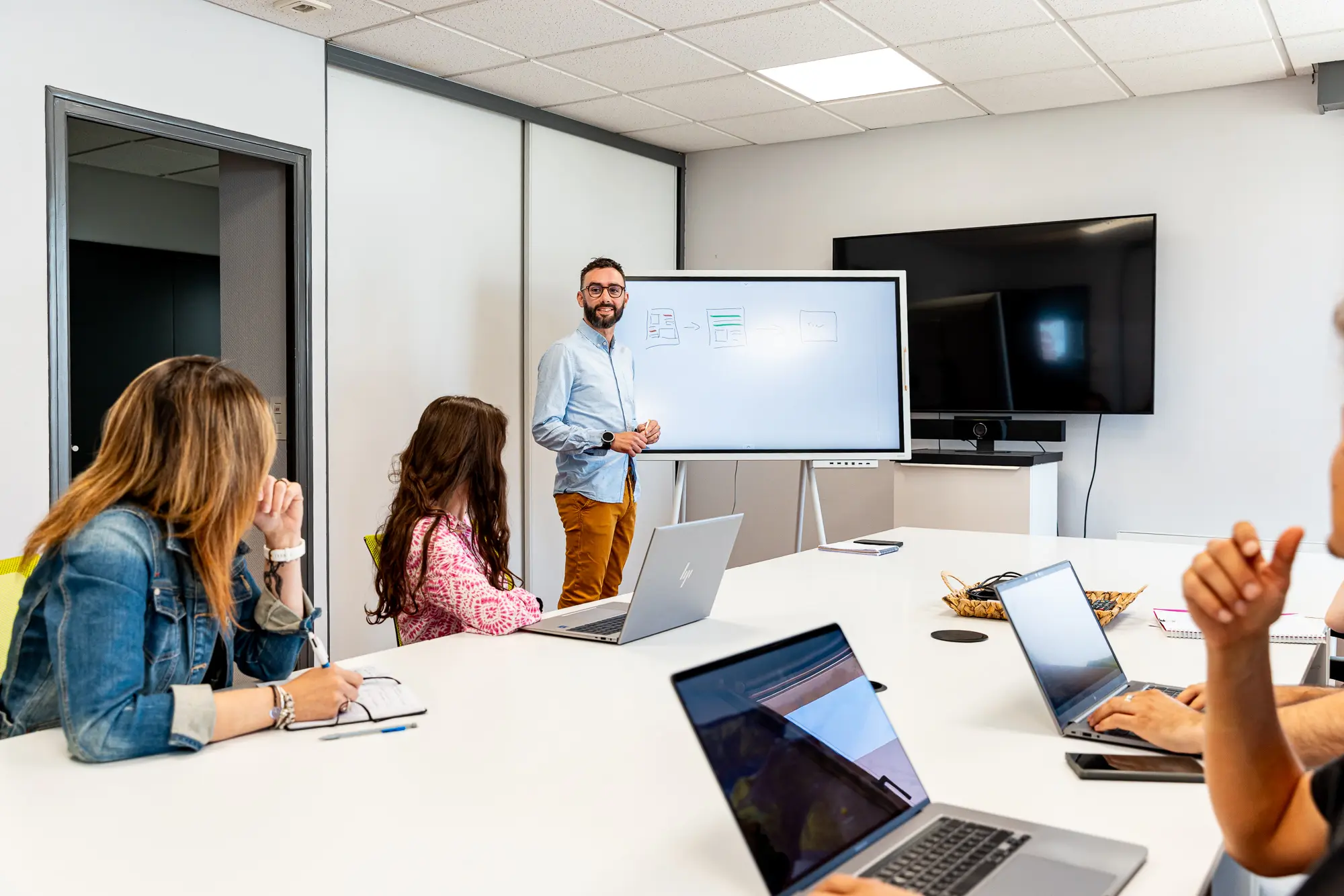 Équipe en réunion dans une salle de conférence, un homme présente sur un tableau blanc et vitrine numérique, plusieurs collègues avec ordinateurs portables. Équipe en réunion dans une salle de conférence, un homme présente sur un tableau blanc et vitrine numérique, plusieurs collègues avec ordinateurs portables.
