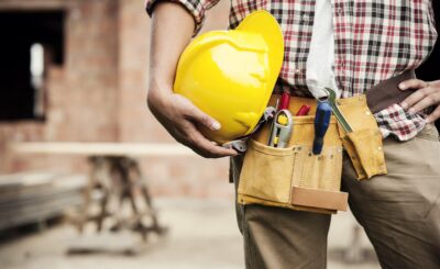 Ouvrier de chantier tenant un casque jaune et une ceinture à outils, prêt pour des travaux de gros œuvre. Cahier des charges gros œuvre sur fond de chantier.