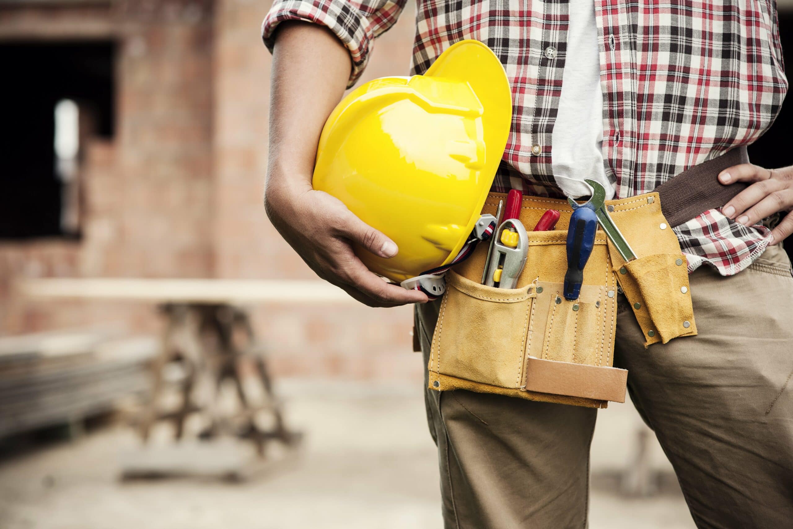 Ouvrier de chantier tenant un casque jaune et une ceinture à outils, prêt pour des travaux de gros œuvre. Cahier des charges gros œuvre sur fond de chantier.
