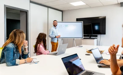 Chef de formation devant un tableau blanc lors d'une réunion, participants avec ordinateurs portables autour d'une grande table. rapport d'analyse des offres, usage en commande publique.