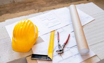 Maître d’ouvrage définition : plans et outils de chantier, casque jaune, règle et coupe-fil sur les plans.