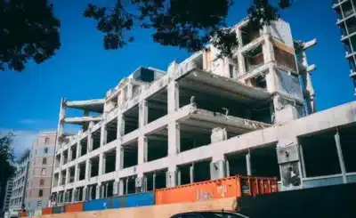 Gratte-ciel en construction ou réhabilitation, structure en béton apparente et grues, containers de chantier colorés au premier plan.